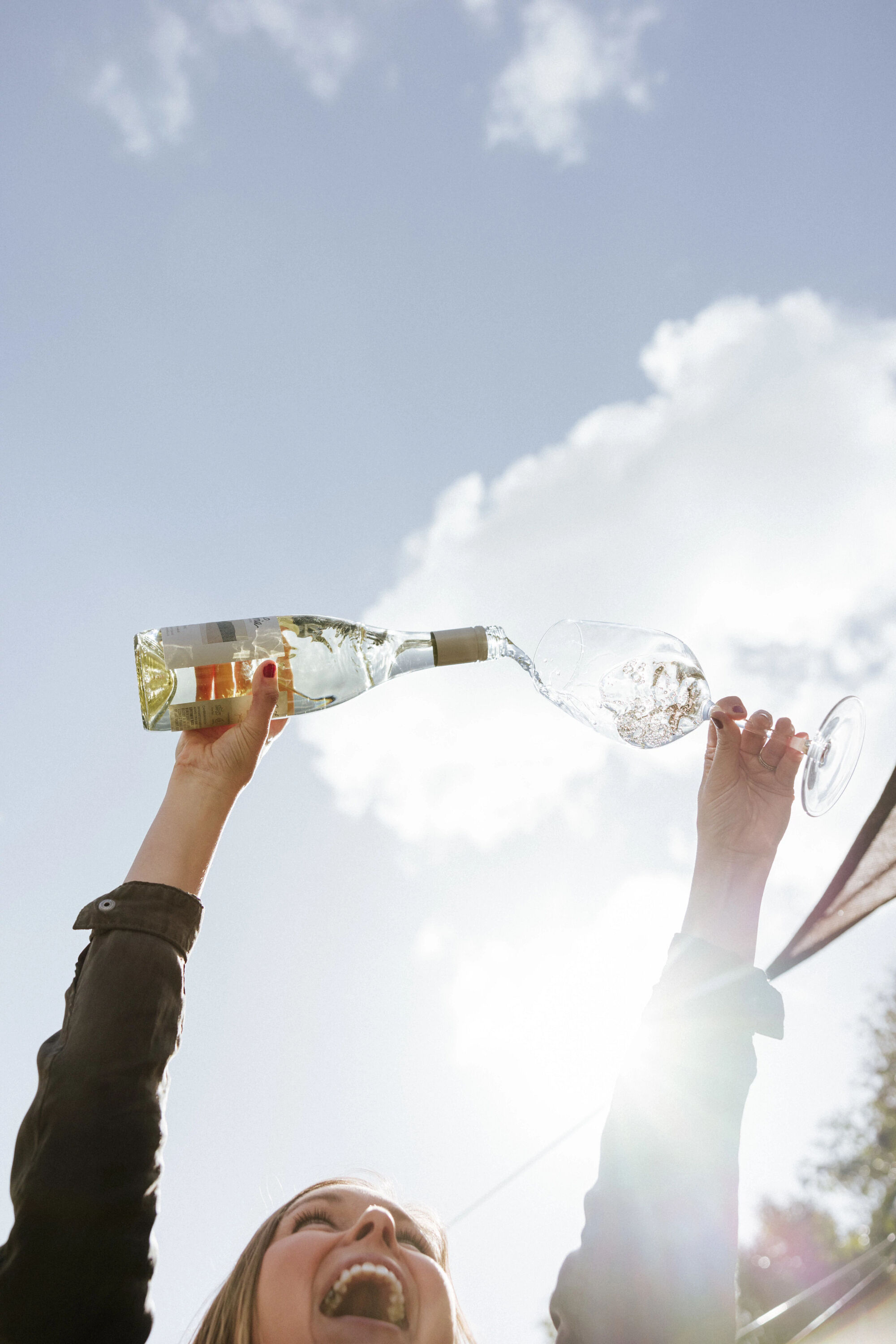 A woman looking up at a bottle of white wine and a glass smiling and pouring against a blue sky