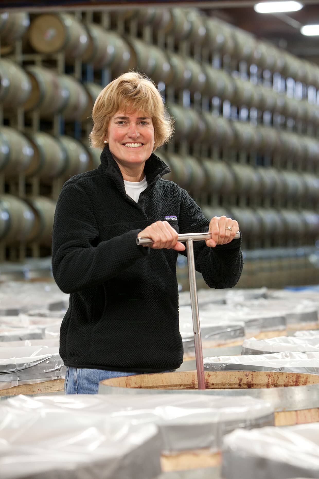 A woman in a black sweatshirt doing a punch down of wine and smiling at the camera