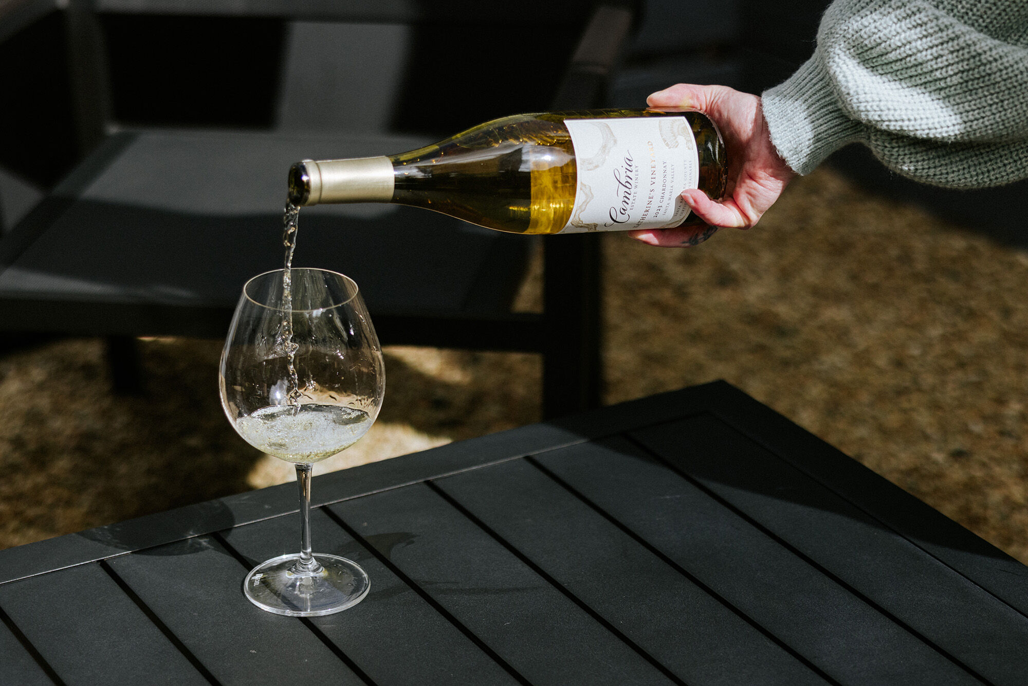 A glass of white wine on a black table outdoors with a person pouring wine into it