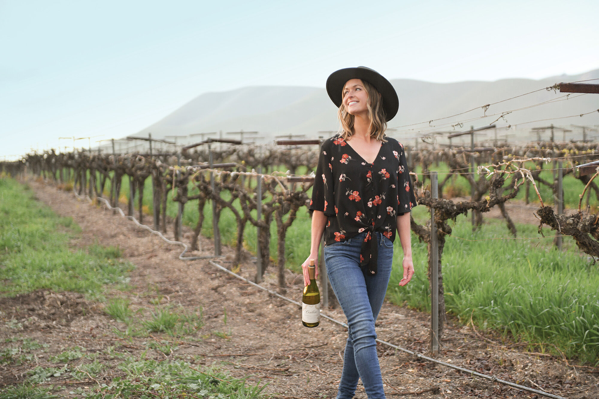 A woman in a black and red shirt, a black hat, and jeans holding a bottle of wine walking through a vineyard