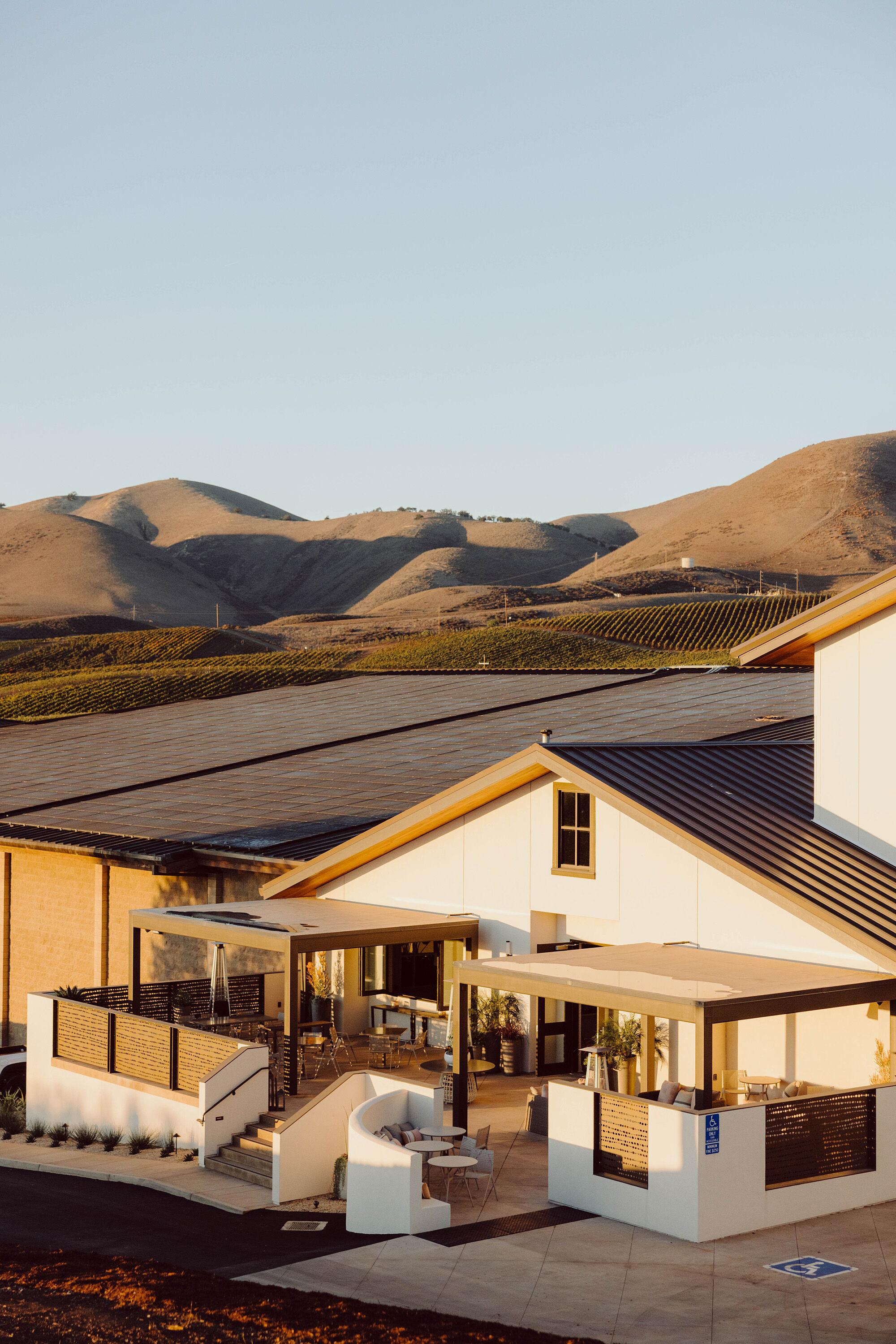 A white and brown winery building with solar panels on the roof and brown hills in the background