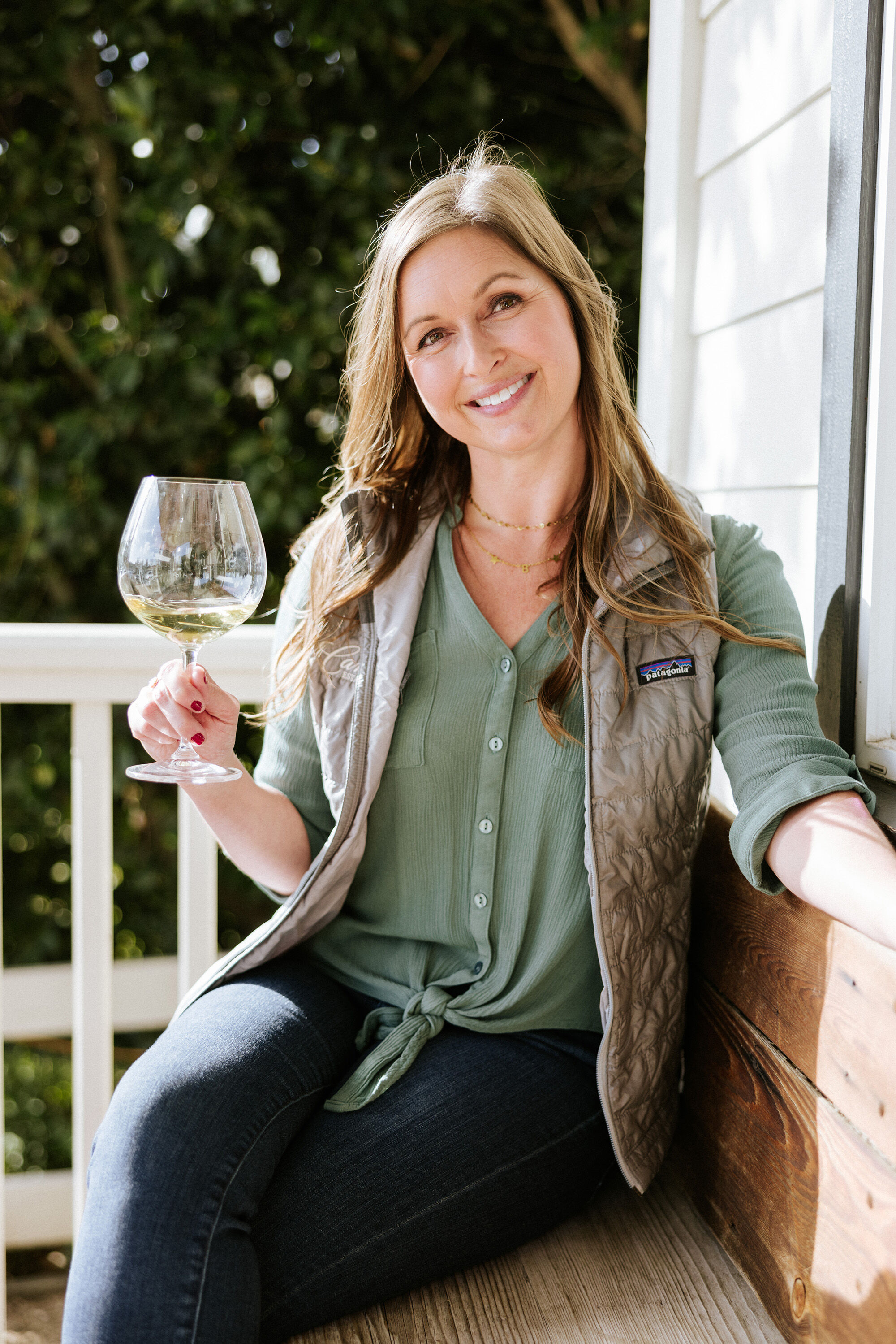 A woman in a green shirt and brown vest smiling with a glass of white wine in her hand