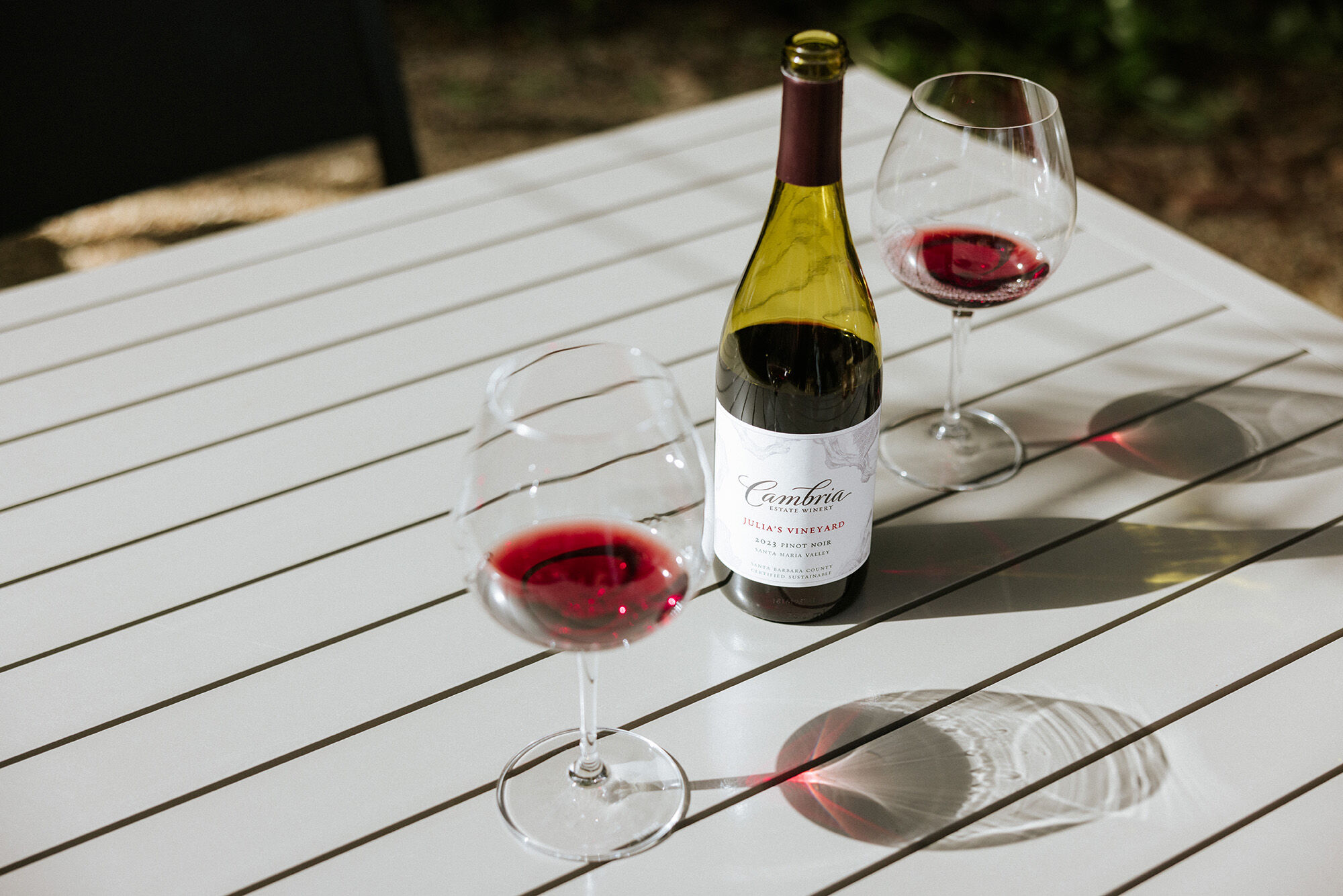 A bottle of red wine in a grey table outdoors with two glasses of red wine next to it on either side